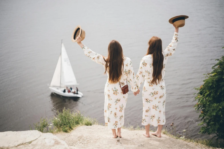 a couple of women standing on top of a cliff next to a body of water, by Julia Pishtar, pexels contest winner, floral clothes, shaped like a yacht, twins, golden-white robes