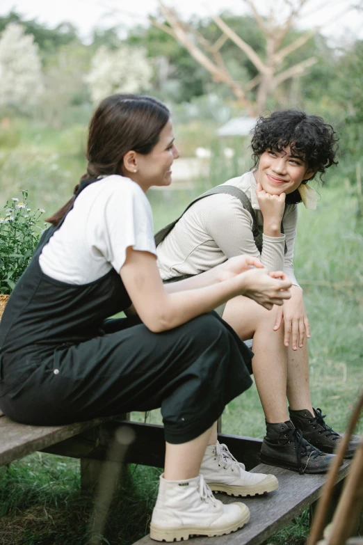 a couple of people that are sitting on a bench, wearing overalls, dark short curly hair smiling, lesbians, trending photo