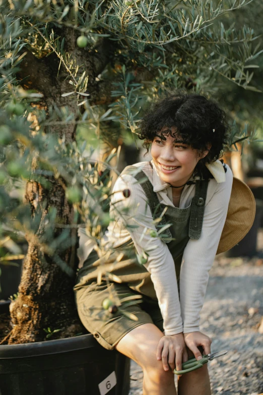 a woman kneeling next to a potted olive tree, trending on pexels, dark short curly hair smiling, wearing farm clothes, avatar image, sophia lillis