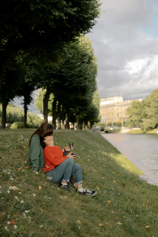 a couple of people sitting on the side of a river, by Anna Boch, pexels contest winner, visual art, saint petersburg, vhs colour photography, beer, with a park in the back ground