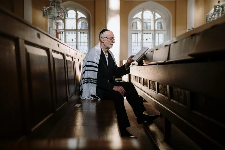 a man sitting on a bench reading a book, by Matthias Stom, unsplash, modernism, mosque synagogue interior, portrait of benjamin netanyahu, standing in front of the altar, full body image