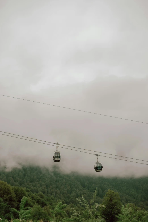 a couple of gondolas sitting on top of a lush green hillside, by Peter Churcher, hurufiyya, overcast!!!, fog!!!, trams ) ) ), suspended in air