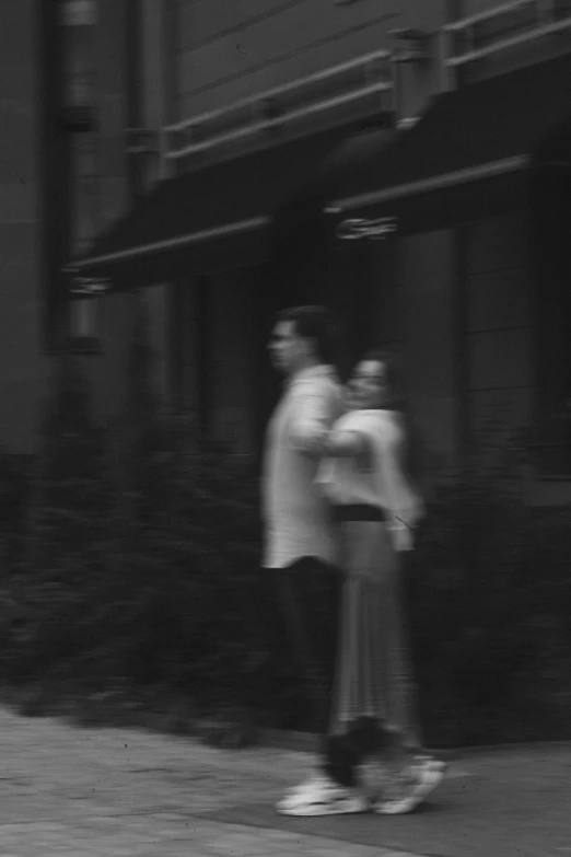 a man riding a skateboard down a sidewalk, a black and white photo, inspired by Garry Winogrand, man and woman walking together, wearing white cloths, low quality footage, haunted