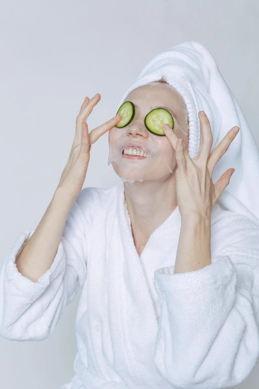 a woman with cucumber slices on her eyes, by Julia Pishtar, shutterstock, renaissance, smiling mask, wearing a grey robe, photoshoot for skincare brand, hands shielding face