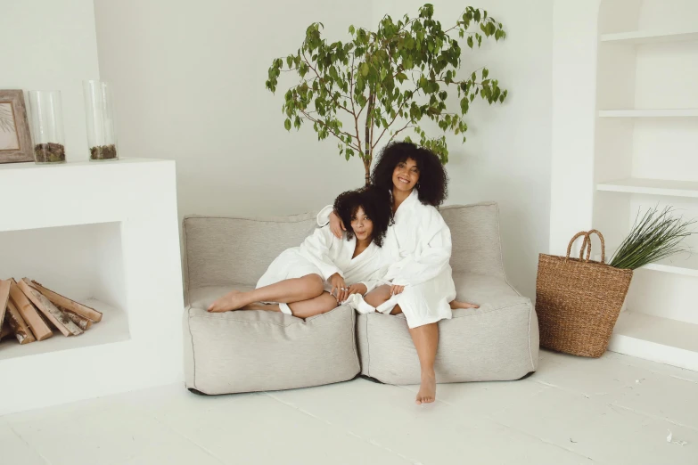 a couple of women sitting on top of a couch, by Emma Andijewska, pexels contest winner, minimalism, bathrobe, botanical, white l shaped couch, teenager hangout spot