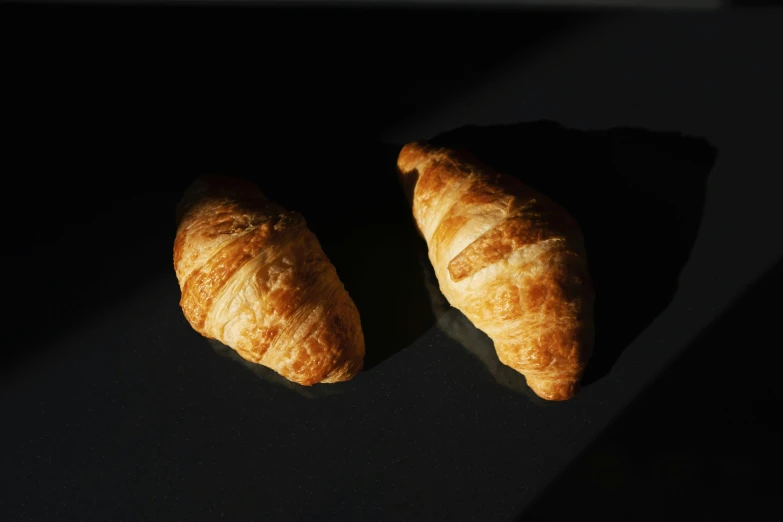 a couple of croissants sitting on top of a table, in front of a black background, in the sun, detailed product image, flattened