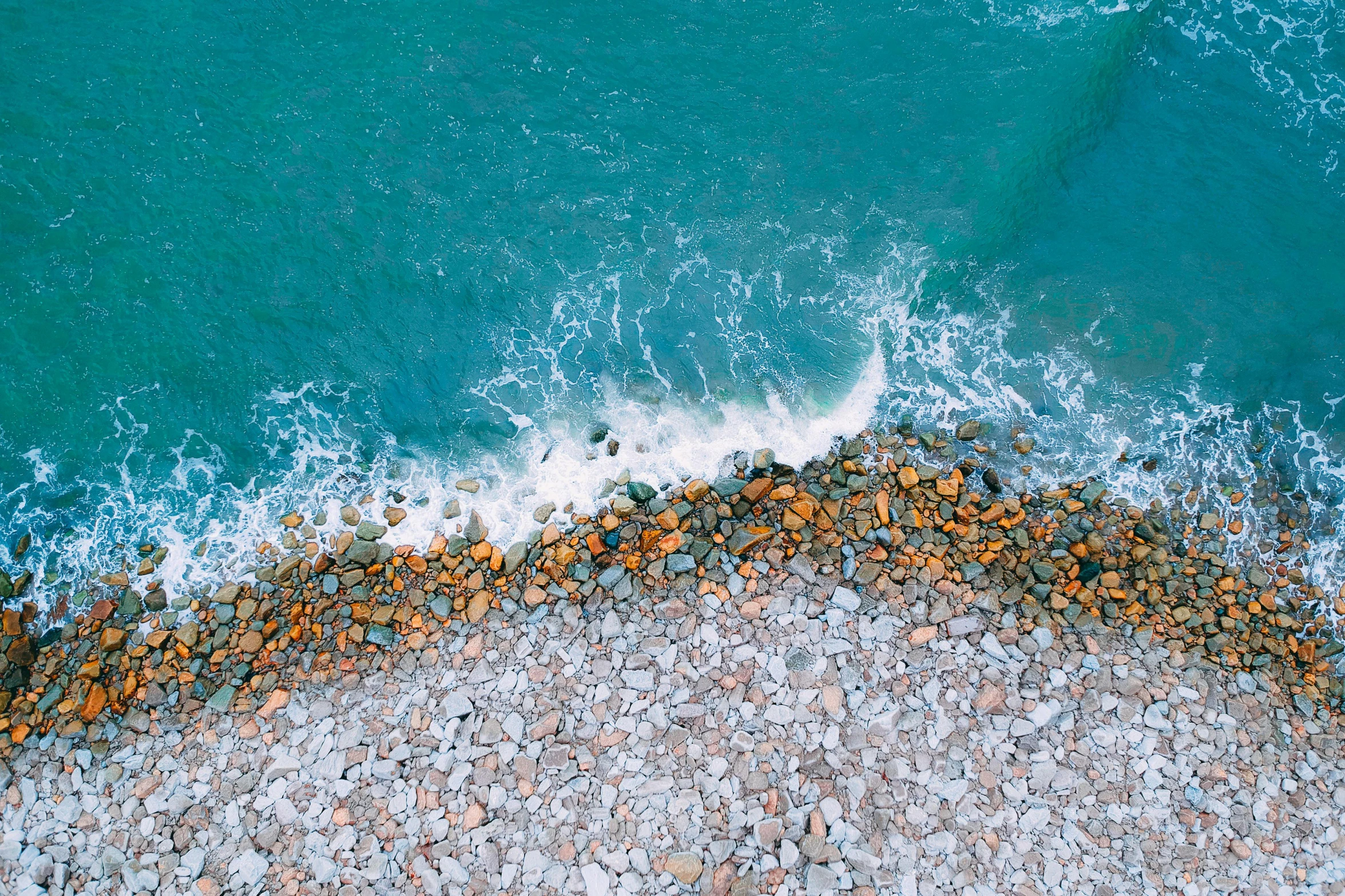 a person riding a surfboard on top of a rocky beach, inspired by Andreas Gursky, pexels contest winner, orange and teal color, birdseye view, sea foam, gravel and scree ground