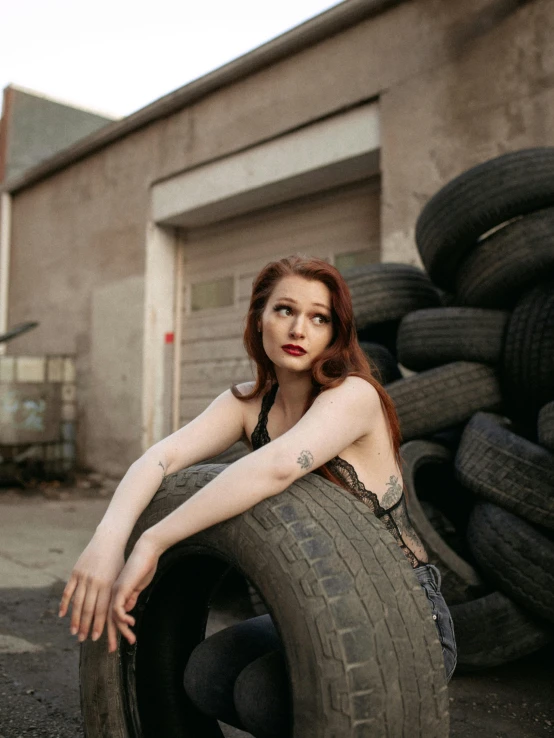 a woman sitting on top of a pile of tires, inspired by Elsa Bleda, trending on pexels, renaissance, red hair and attractive features, in front of a garage, non binary model, with pale skin