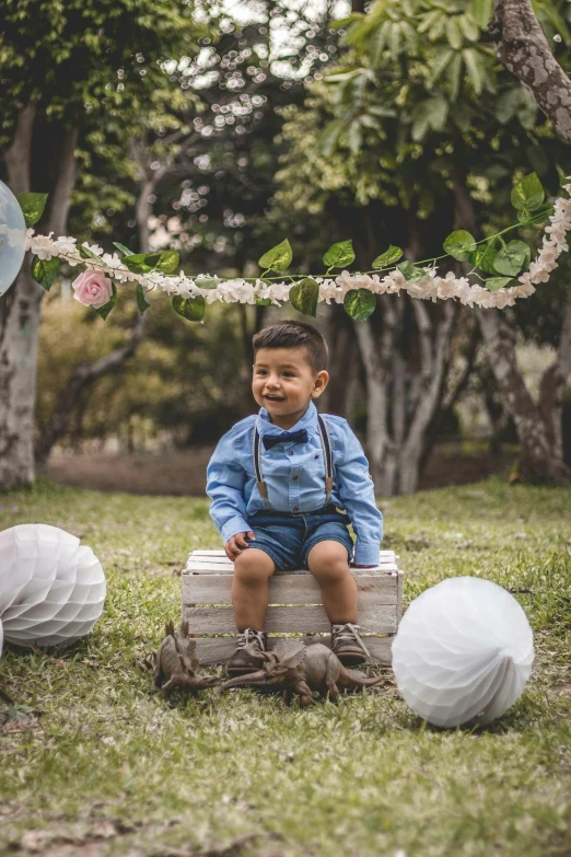 a little boy sitting on top of a wooden box, with paper lanterns, wearing blue jean overalls, zigor samaniego, with flowers