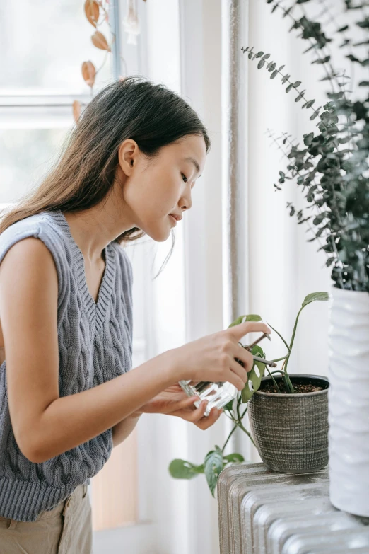 a woman standing next to a radiator holding a plant, pexels contest winner, checking her phone, asian woman, plants in beakers, carefully crafted