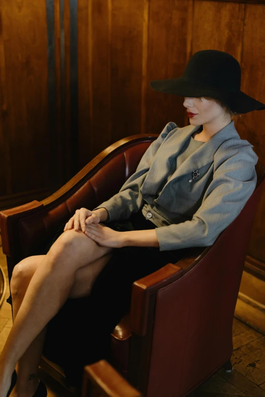 a woman sitting in a chair in a room, inspired by Nan Goldin, unsplash, renaissance, a suited man in a hat, met collection, 1930s style clothing, grey jacket