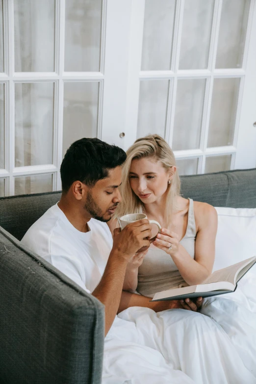 a man and woman sitting on a bed looking at a laptop, by Cosmo Alexander, pexels contest winner, romanticism, white mug, reading for a party, sitting on couch, next to a cup