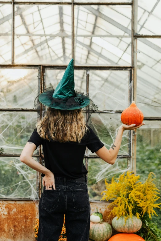 a woman in a witches hat holding a pumpkin, by Julia Pishtar, pexels contest winner, glass greenhouse, beistle halloween decor, inspect in inventory image, back - shot