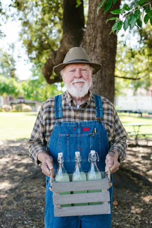 a man in overalls holding a box of bottles, a portrait, by Leland Bell, pexels, white hair and white beard, parks, california;, promo image