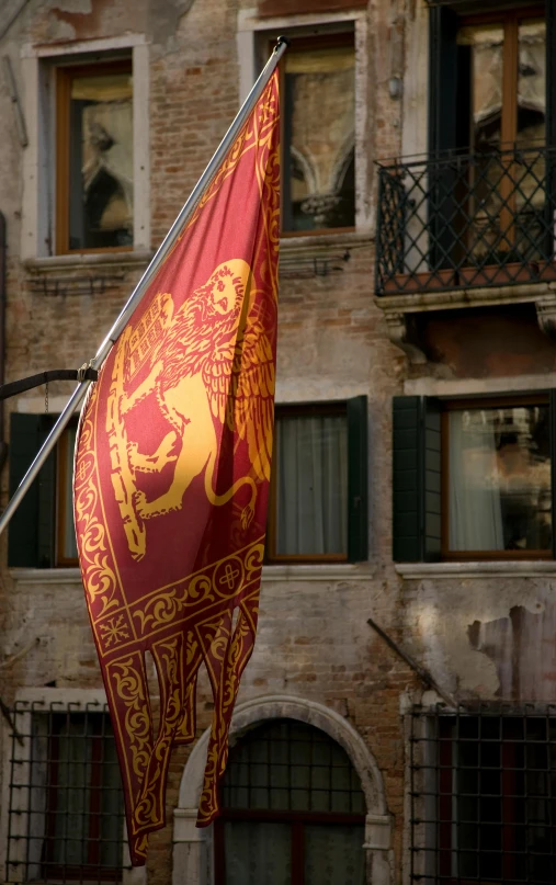 a red and gold flag on a pole in front of a building, inspired by Antonio de la Gandara, renaissance, venice biennale's golden lion, maroon and white, medieval setting, square