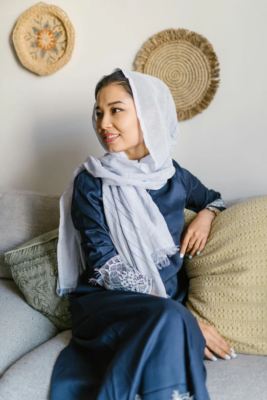 a woman sitting on a couch in a living room, inspired by Maryam Hashemi, hurufiyya, dress in voile, white and blue, white sleeves, white scarf