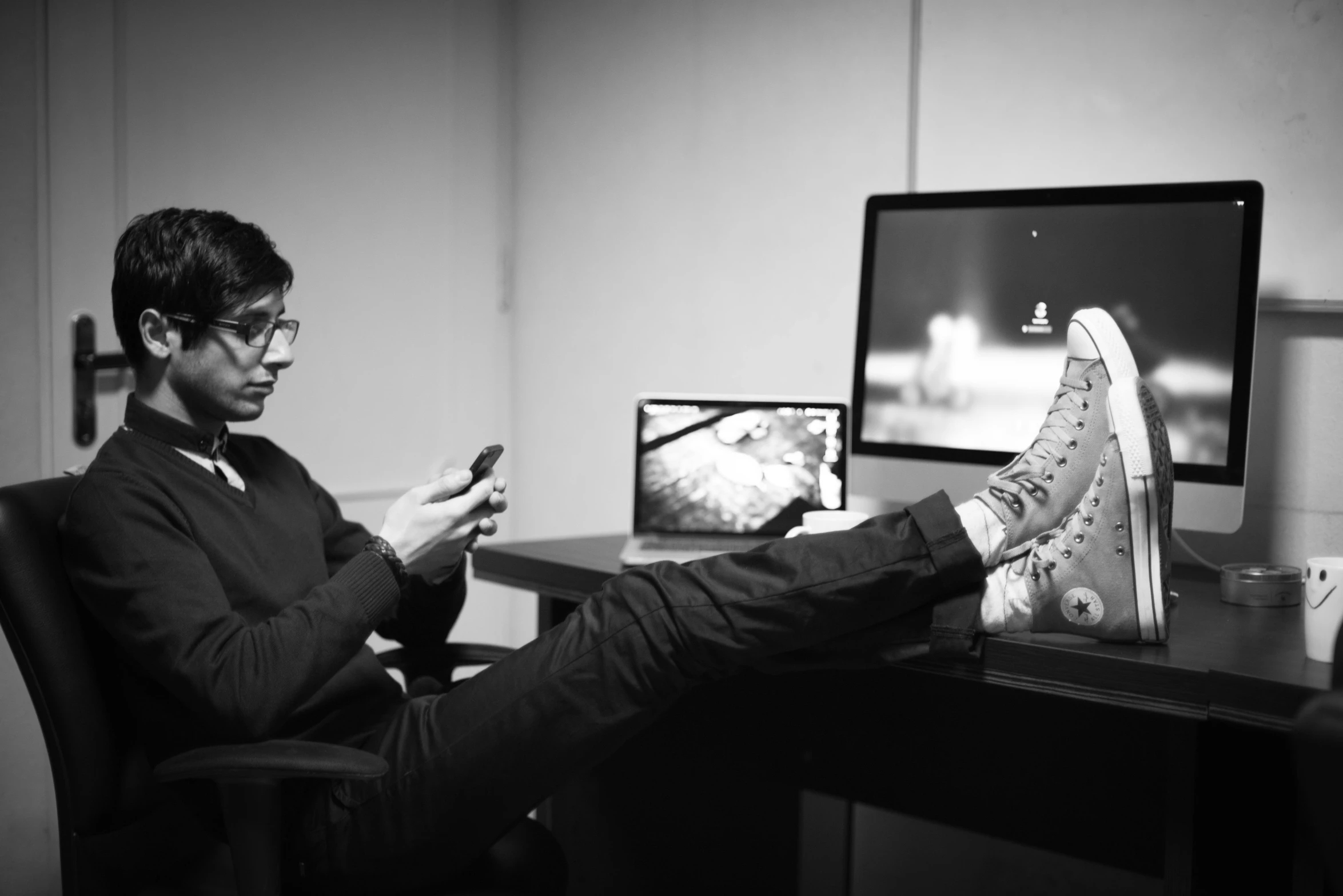 a man sitting at a desk using a cell phone, a black and white photo, by Romain brook, wearing boots, mia khalifa, kent monkman, monitor