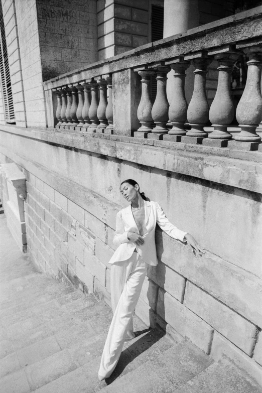 a black and white photo of a woman leaning against a wall, inspired by Helmut Newton, unsplash, wearing white suit, in balcony of palace, lulu chen, ffffound