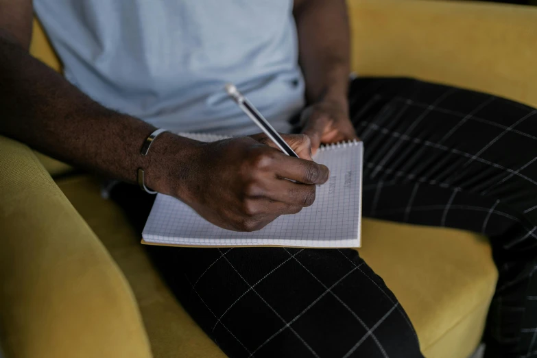 a man sitting on a couch writing in a notebook, by Carey Morris, pexels contest winner, man is with black skin, on a checkered floor, from waist up, without text