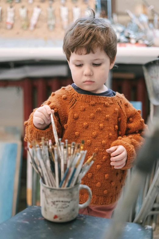 a little boy standing in front of a pot full of paint brushes, inspired by Louis Le Nain, pexels contest winner, he is wearing a brown sweater, carefully crafted, organic detail, lights on