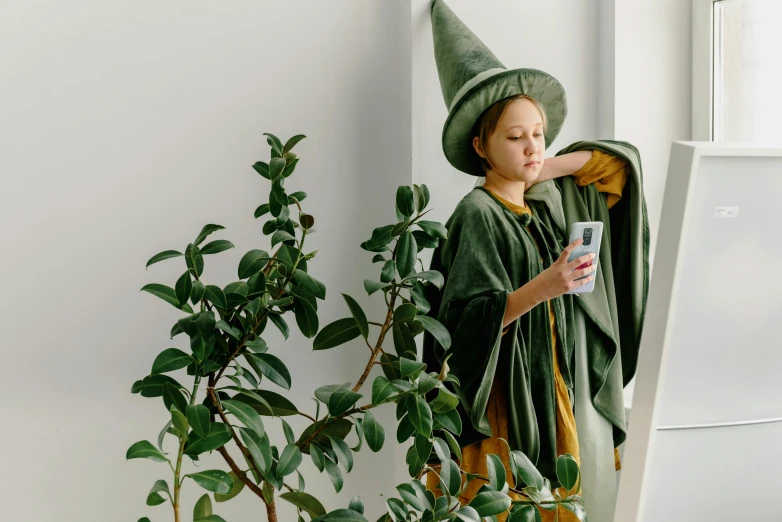 a little girl standing next to a potted plant, inspired by Elsa Beskow, trending on pexels, magical realism, wearing a wizard cloak, looking at his phone, avacado halloween costumes, green hat