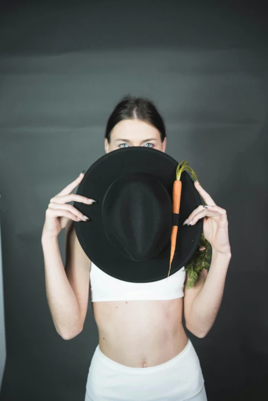 a woman holding a black hat over her face, an album cover, inspired by Marina Abramović, unsplash, antipodeans, carrots, bare midriff, studio shoot, veggies