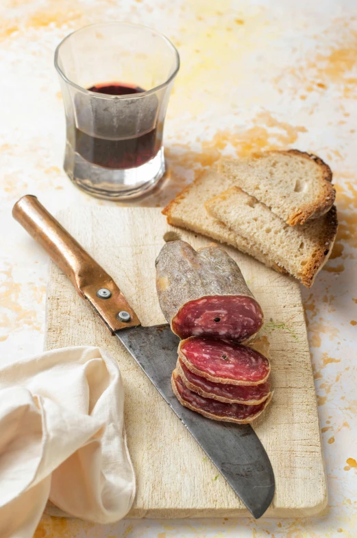 a knife sitting on top of a cutting board next to a glass of wine, brown bread with sliced salo, salami, indigo