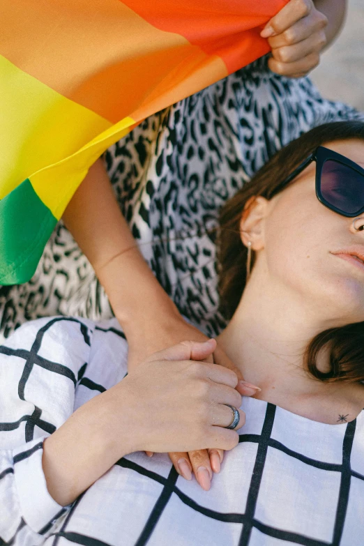 a woman laying on the ground with a rainbow flag, by Julia Pishtar, trending on unsplash, with sunglass, woman holding another woman, laying on a bed, furr covering her chest