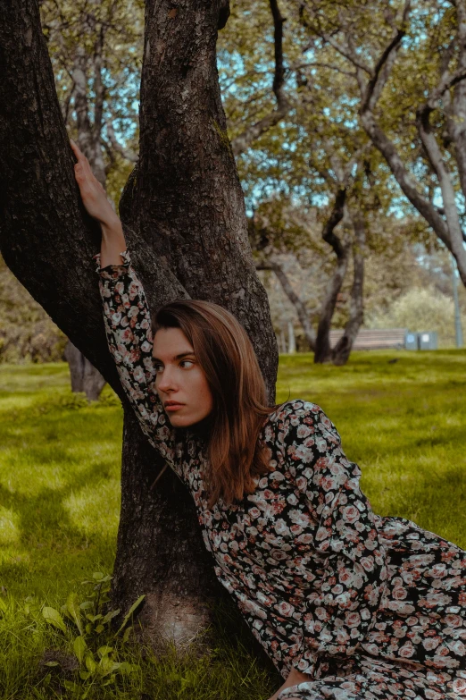 a woman leaning against a tree in a park, a colorized photo, inspired by Elsa Bleda, pexels contest winner, arabesque, trees and flowers, girl with brown hair, doing a sassy pose, patterned clothing