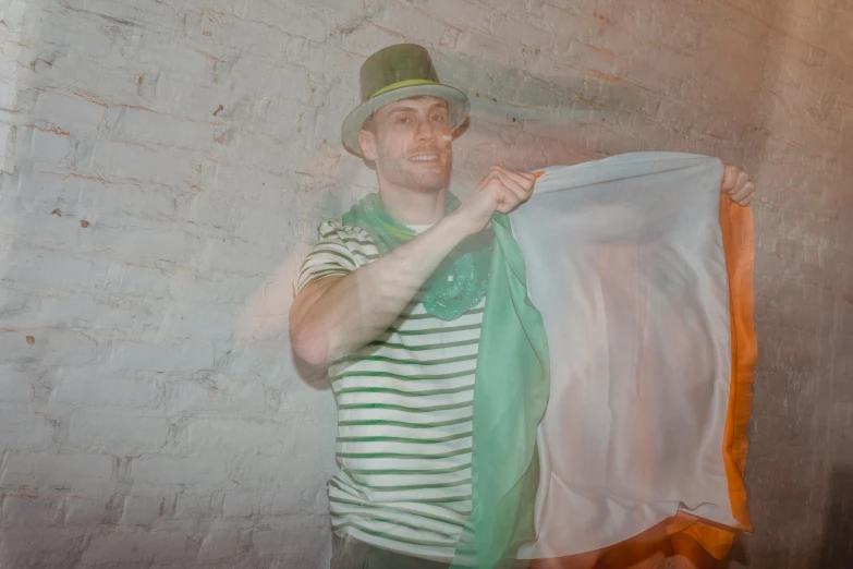 a man standing in front of a brick wall holding a green and white scarf, by Anna Füssli, happening, wearing green tophat, dj at a party, covered in transparent cloth, magic experiment