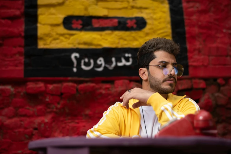 a man sitting at a table in front of a brick wall, an album cover, by Ahmed Yacoubi, trending on pexels, graffiti, egyptian, king in yellow, movie still frame, background image