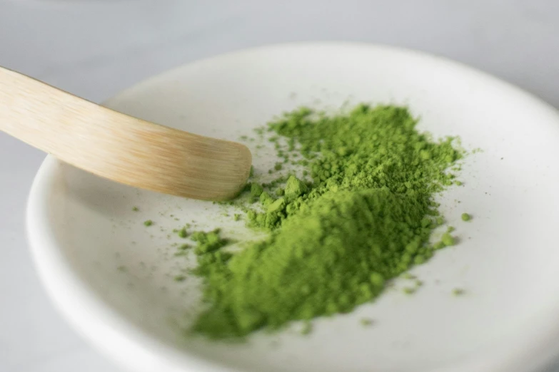 a white bowl filled with green powder and a wooden spoon, inspired by Masamitsu Ōta, hurufiyya, press shot, grassy, medium close up, lifestyle