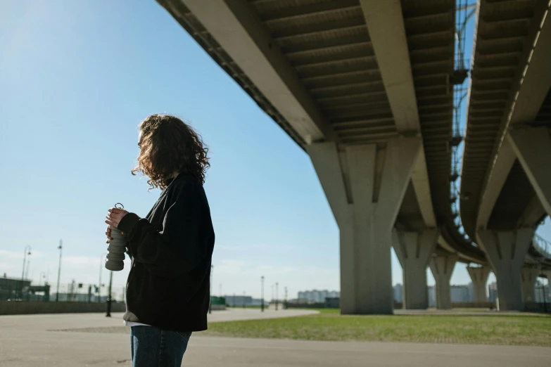 a woman standing under a bridge holding a cup of coffee, unsplash contest winner, happening, film still from the movie, city bay bridge aqueduct, holding a bottle, contemplating