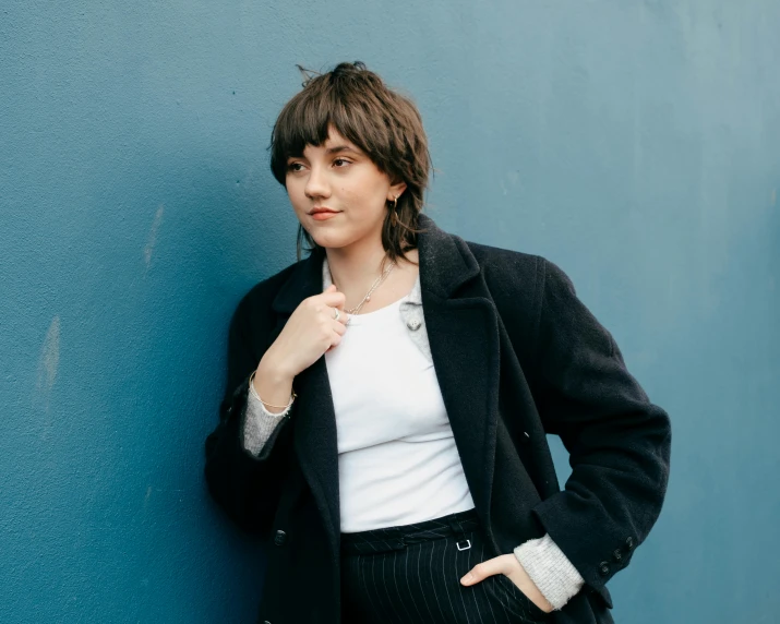 a woman leaning against a blue wall, an album cover, inspired by Elsa Bleda, pexels contest winner, realism, maisie williams, a man wearing a black jacket, curly bangs, kaitlyn michelle siragusa