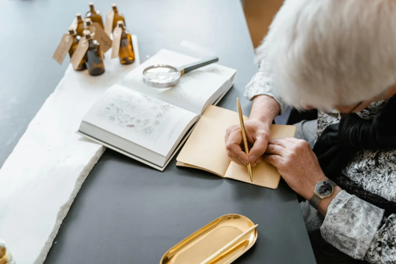 a woman sitting at a table writing on a piece of paper, by Matthias Stom, pexels contest winner, silver gold details, thumbnail, flatlay book collection, grandma