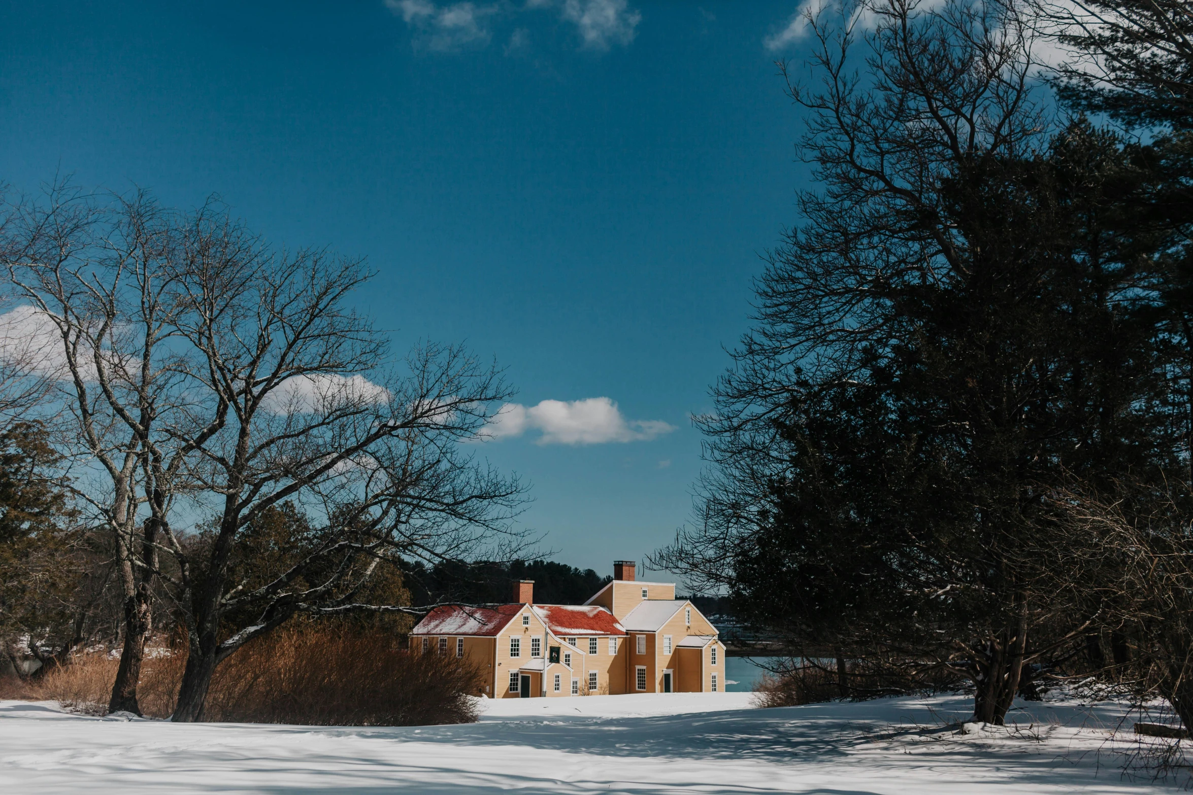 a house sitting in the middle of a snow covered field, by Jessie Algie, pexels contest winner, colonial style, wes anderson background, lakeside, sunny day time