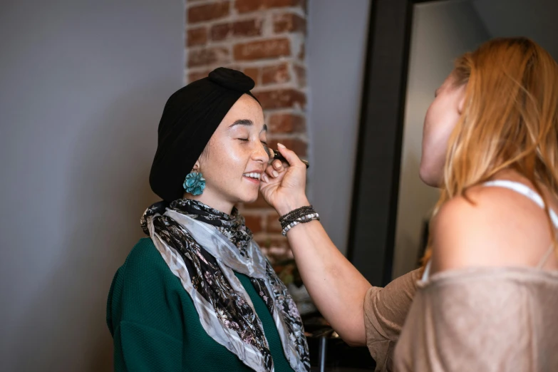 a woman putting makeup on another woman's face, hurufiyya, fan favorite, wearing a head scarf, in professional makeup, gemma chen
