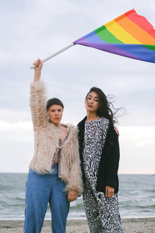 two women holding a rainbow flag on a beach, an album cover, trending on pexels, wearing a fancy jacket, confident pose, 🚿🗝📝, androgynous male