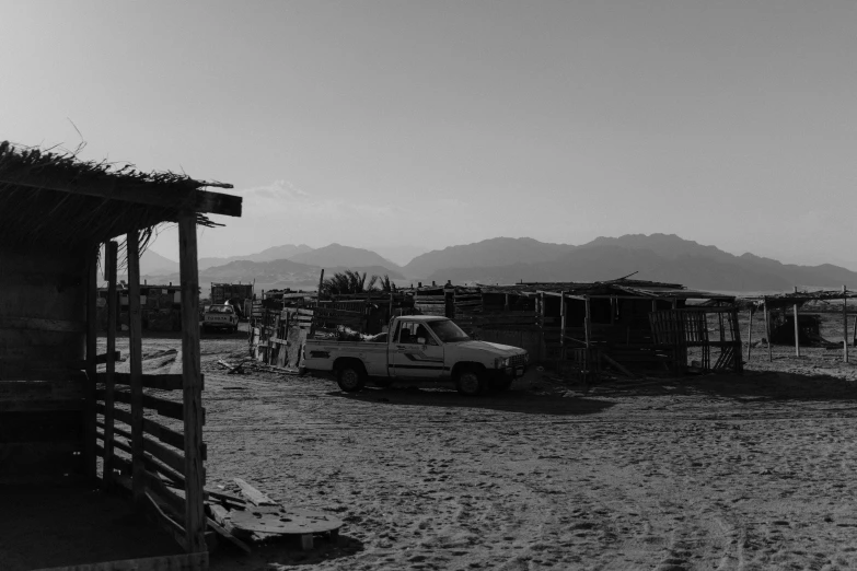 a black and white photo of a truck parked in front of a shack, a black and white photo, unsplash, mexican desert, background image, some stalls, with mountains in the background