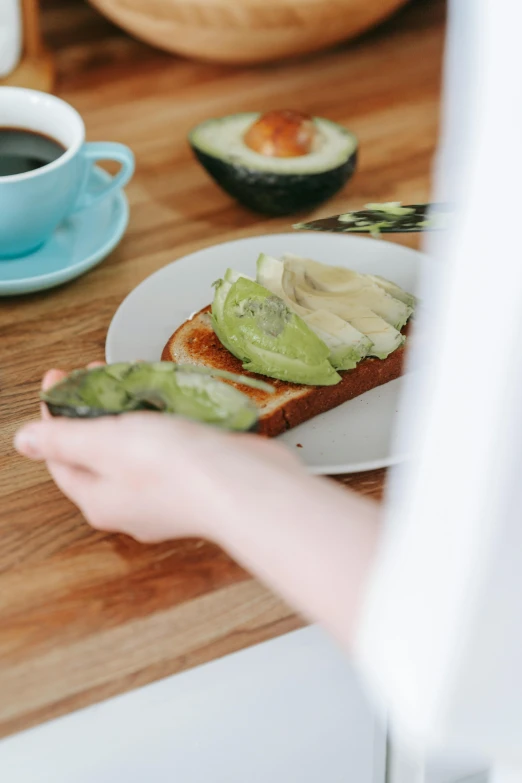 a person holding a piece of bread with avocado on it, pexels contest winner, ☕ on the table, summer morning, jen atkin, promo image