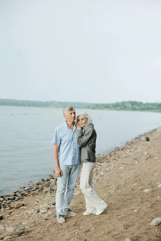 a man and a woman standing next to a body of water, gray haired, lifestyle, rocky lake shore, medium - shot