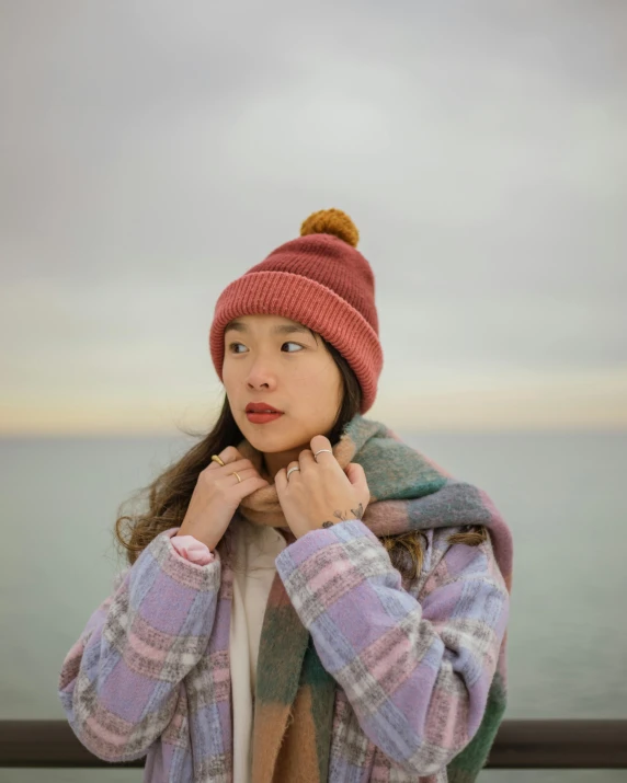 a woman standing in front of a body of water, a colorized photo, by helen huang, trending on pexels, wearing beanie, maroon, young cute wan asian face, model wears a puffer jacket