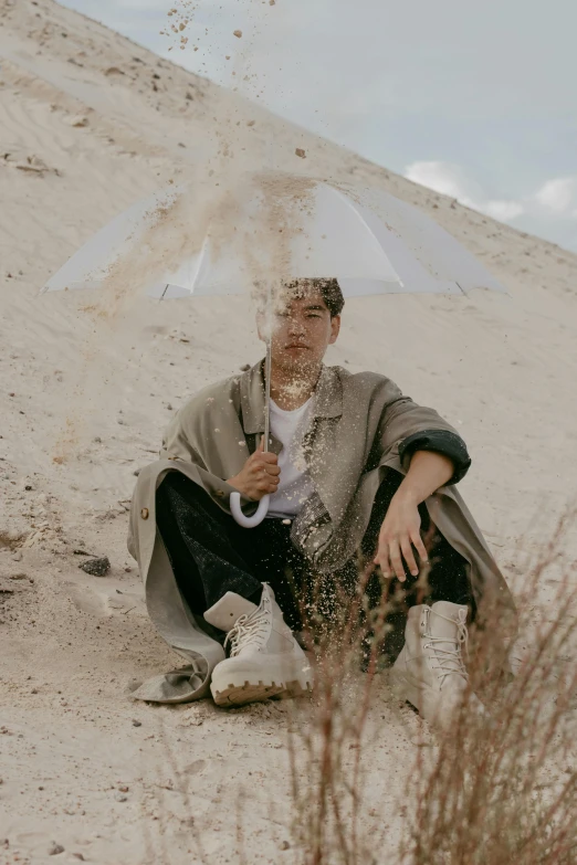 a man sitting in the sand with an umbrella, bad bunny, leslie zhang, wearing dirty travelling clothes, full of sand and glitter