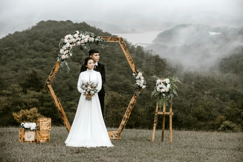 a bride and groom standing in front of a wedding arch, pexels contest winner, foggy forrest backdrop, in style of lam manh, hills in the background, bouquets