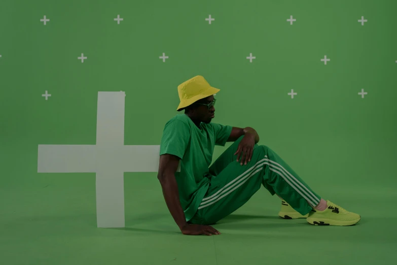 a man sitting on the ground in front of a cross, trending on pexels, video art, green hat, wearing adidas clothing, ( ( dark skin ) ), yellow and green