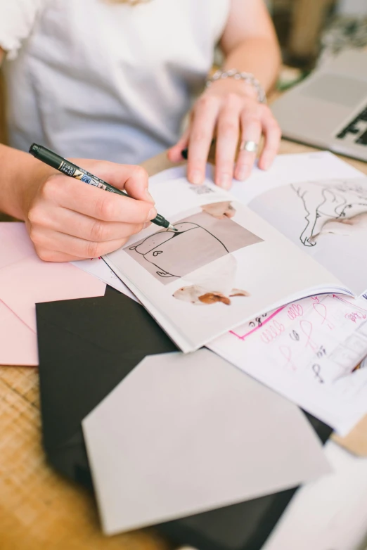 a woman sitting at a table writing on a piece of paper, a drawing, clothing design, 9 9 designs, pink and grey muted colors, holding notebook