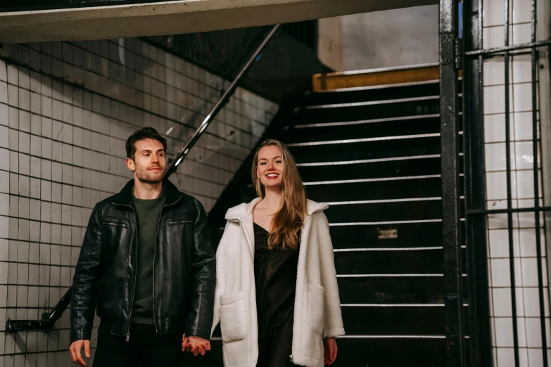 a man and a woman standing next to each other, a photo, by Emma Andijewska, pexels contest winner, subways, wearing an aviator jacket, promotional image, holding hands