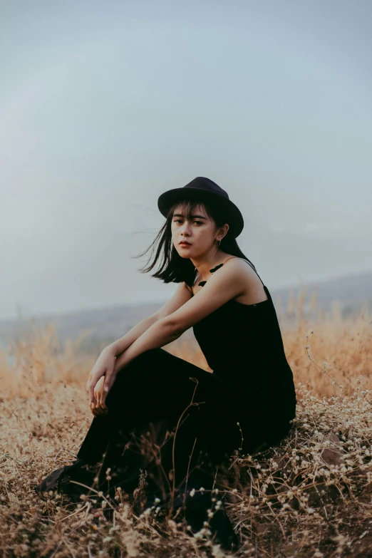 a woman sitting on top of a grass covered field, an album cover, trending on pexels, realism, black hat, asian girl, portrait mode photo, thinking pose