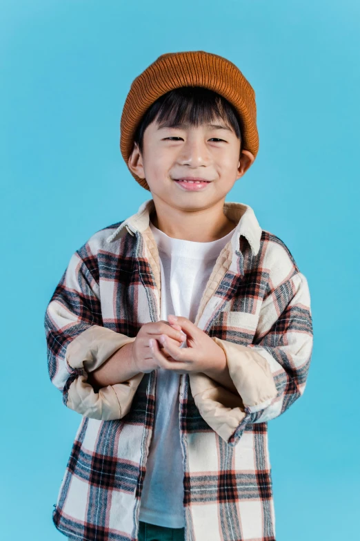 a young boy standing in front of a blue background, trending on pexels, mingei, wearing a flannel shirt, caracter with brown hat, asian human, a still of a happy