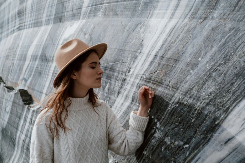 a woman in a hat leaning against a wall, a marble sculpture, inspired by Andrew Wyeth, trending on pexels, geological strata, wearing a sweater, structural geology, grey